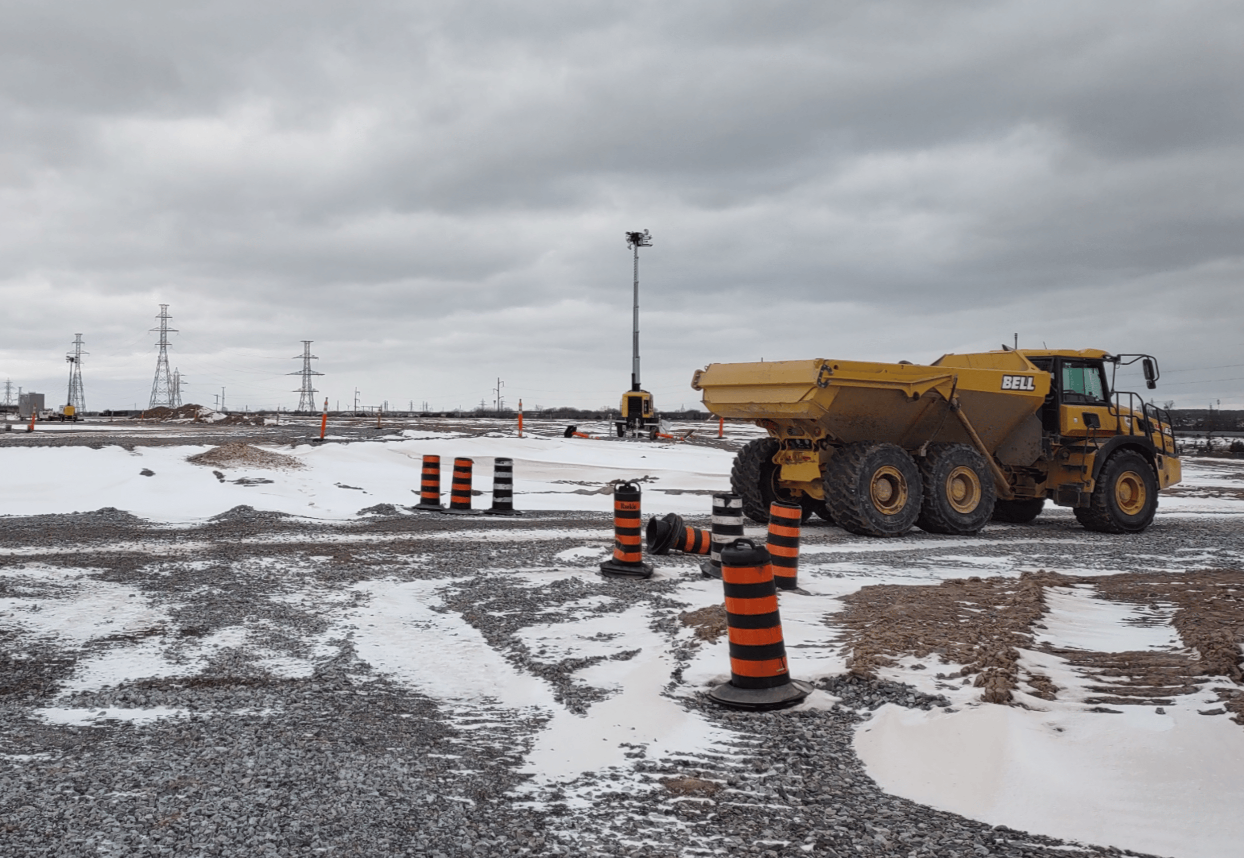 Articulated dump truck operating on a winter construction jobsite with temporary traffic barrels and a mobile light tower